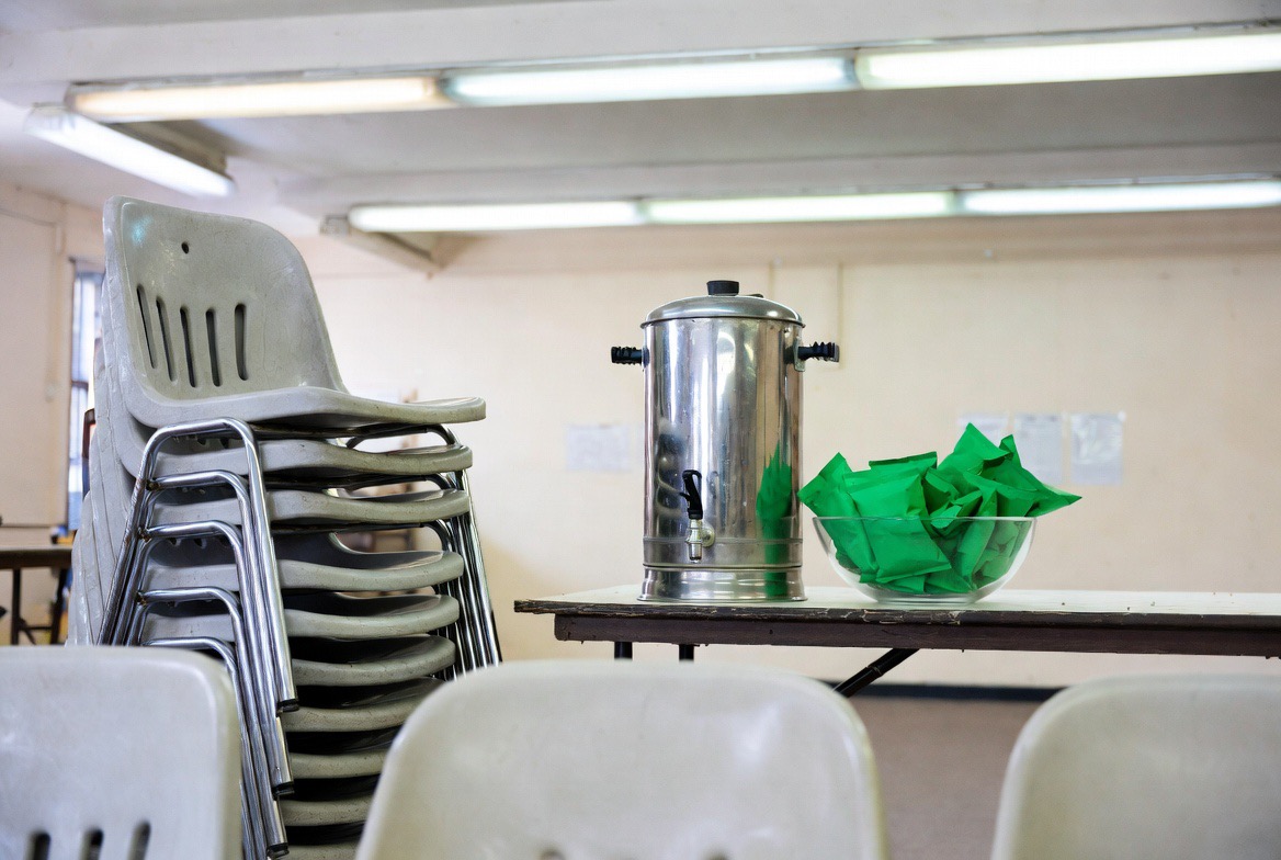 A community hall under fluorescent lights: a stack of plastic chairs beside a folding table holding a metal tea urn and a glass bowl filled with green crisp packets.